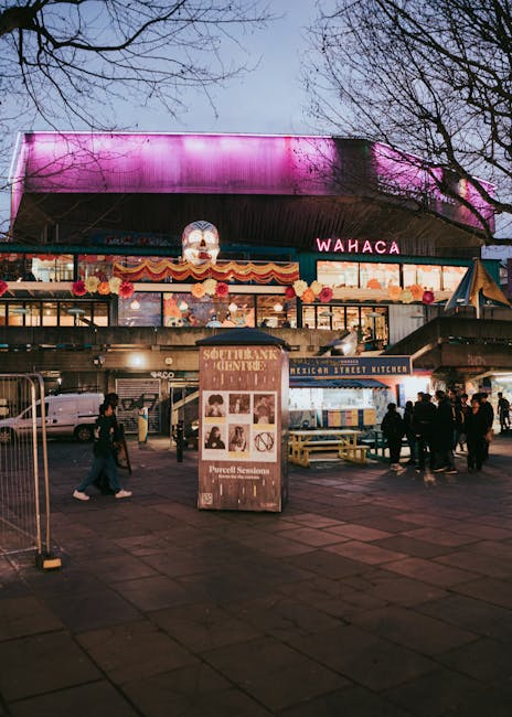 The image depicts a lively urban scene in the early evening or dusk, with a large, modern building featuring a curved roof illuminated by pink and purple lighting. In the foreground, there is a paved area with several pedestrians walking and gathering around food stalls and outdoor seating, which include wooden picnic tables. The food stalls have bright signage, including one labeled 'Mexican Street Kitchen,' and a prominent display board in front advertising 'Southbank Centre.' Surrounding the scene are leafless trees, suggesting late autumn or winter, and parked cars are visible behind the pedestrian area. The overall atmosphere combines metropolitan vibrancy with outdoor social spaces, and although the focus is on a commercial or entertainment setting, the scene reflects well-maintained pathways, organized seating, and an inviting environment, typical of a lively, landscaped urban area. This image aligns with themes of city landscaping and outdoor community spaces, as seen in the Waterloo gardeners guide to small South Bank terraces, supported by Gardeners Waterloo.
